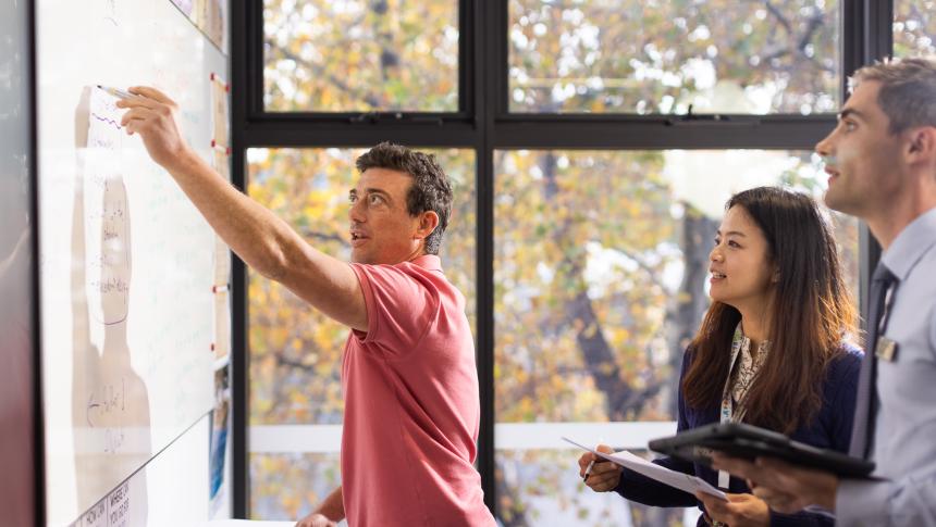 Three adults writing notes on whiteboard in a classroom.