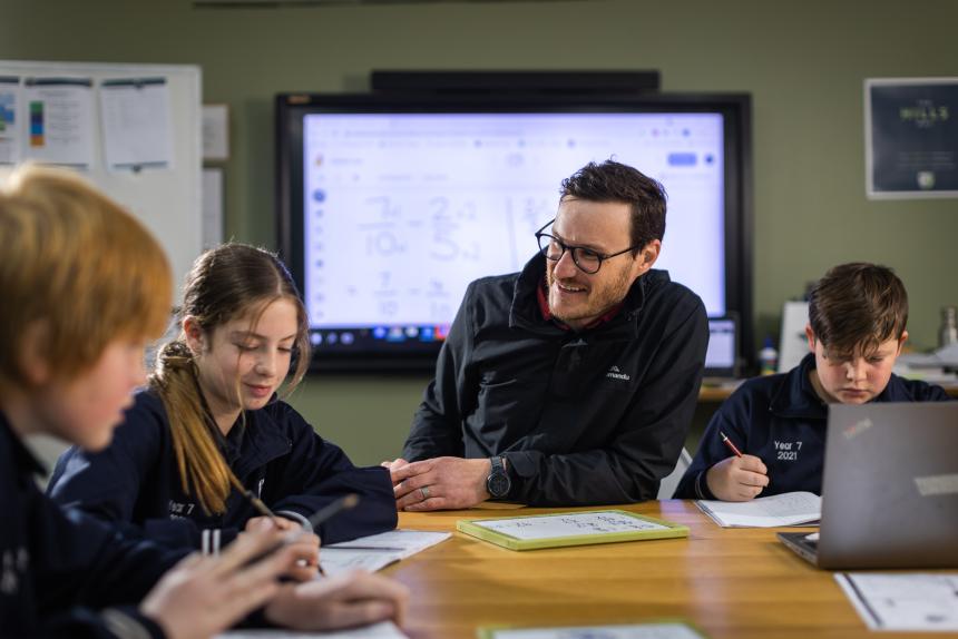 A secondary classroom setting with a male teacher sitting at a table with three students. They hold pens with paper and laptops in front of them. 