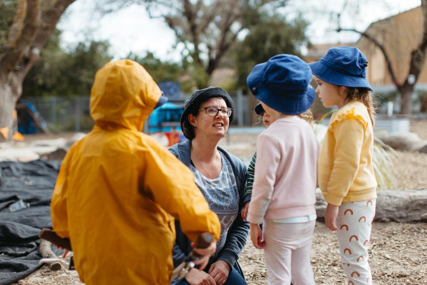An early childhood educator outside with three children. The educator is kneeling down to speak to them at their level. 