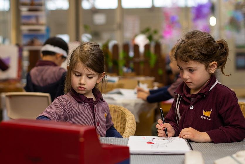 Two young children, about 4 or 5 years old, sitting at a desk with a pen and book 