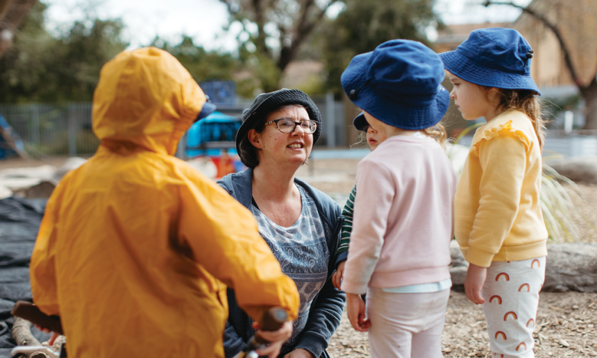 An early childhood educator outside with three children. The educator is kneeling down to speak to them at their level. 