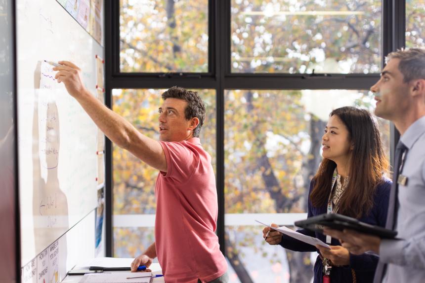 Three adults writing notes on whiteboard in a classroom.